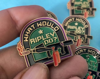 May include: Gold-colored enamel pin with a black and green background. The pin features a cartoon image of a gun with a flame and the text "What Would Ripley Do?" in a white circle with a gold star.