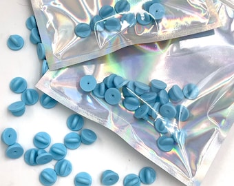 May include: A close-up of a pile of light blue metal pin backs. The pin backs are in a clear plastic bag. The pin backs are used for making custom pins.