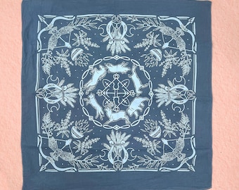 May include: Adriatic Blue bandana with a white, intricate design featuring deer, rabbits, bows, arrows, and other botanical elements. The text "Adriatic Blue" is printed at the bottom. The square cloth is displayed on a pink background.