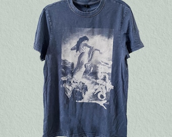 May include: A blue t-shirt featuring a white and gray graphic print of a classical painting. The artwork depicts a woman in flowing robes, with a dramatic, ethereal scene. The shirt has a crew neck and short sleeves.