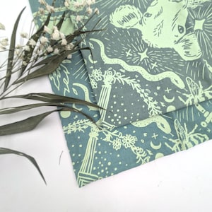 May include: Two teal fabric pieces with a light green celestial design featuring a cow, snake, stars, and floral elements. Dried flowers and leaves are in the foreground, adding a natural touch to the composition. The fabric has a detailed, artistic print.