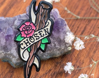 May include: A black enamel pin with a rose, green leaves, and the word "CHOSEN" on a banner. The pin features brown and cream-colored accents, and is set against a purple crystal.