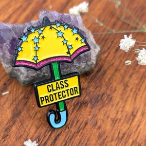 May include: A yellow umbrella-shaped enamel pin with blue star accents and a pink glitter border. The pin has a green stem and a black rectangle with the words "CLASS PROTECTOR" in white. The pin is set on a wooden surface.