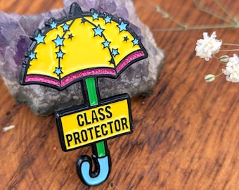 May include: A yellow umbrella-shaped enamel pin with blue star accents and a pink glitter border. The pin has a green stem and a black rectangle with the words "CLASS PROTECTOR" in white. The pin is set on a wooden surface.