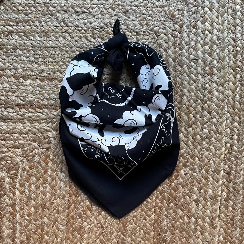 Puede incluir: Bandana negra con un dise&ntilde;o blanco de nubes y gatos. La bandana tiene un nudo en la parte superior.