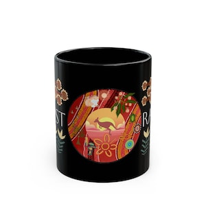 May include: A black ceramic mug featuring a central circular graphic with a kangaroo silhouette against a sunset landscape. The design incorporates dot art patterns in red, orange, and yellow, with floral motifs and partial text visible on the sides.