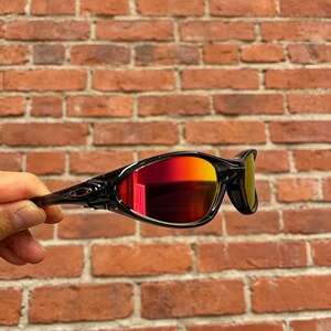 May include: Black wrap-around sunglasses featuring reflective lenses with a vibrant red and orange gradient tint. The frames have a sleek, aerodynamic design with a prominent logo detail on the temple hinge, providing full eye coverage.