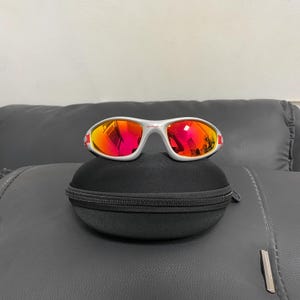 Pode incluir: Óculos de sol prateados com um design de armação envolvente e lentes refletoras em tons vibrantes de vermelho e laranja. Os óculos estão posicionados sobre um estojo de proteção preto, semirrígido, arredondado e com fecho.