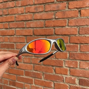 Peut inclure: Lunettes de soleil argentées dotées d'une monture enveloppante avec des verres réfléchissants rouges et orange. Les branches se terminent par des embouts noirs et la monture présente une finition métallique mate avec un logo sur la charnière.