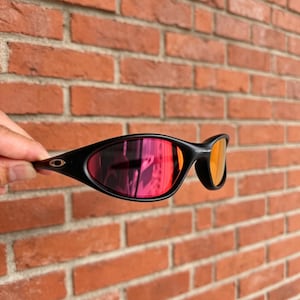 May include: Black sunglasses featuring a wrap-around frame design and reflective lenses that display a vibrant red and orange gradient tint. The temple arm includes a small silver logo detail on the side of the matte black plastic frame.