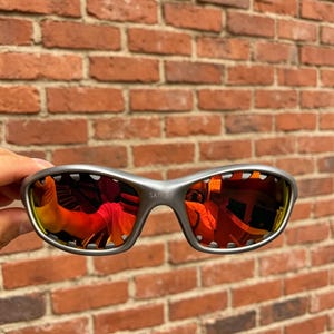 Pode incluir: Óculos de sol em tom prateado com um design de armação envolvente e lentes refletoras laranja e vermelhas. A ponte da armação apresenta a palavra SATIN em letras pequenas em relevo. As lentes mostram um reflexo distorcido do ambiente.