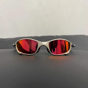 Peut inclure: Lunettes de soleil avec une monture en métal argenté et des verres miroirs rouge vif. La monture présente une construction segmentée distincte avec des rivets visibles au niveau du pont et des charnières, offrant un aspect structuré.