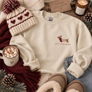 May include: A cream-colored crewneck sweatshirt featuring an embroidered dachshund wearing a winter hat and sweater, with the text LONG STORY SHORT below it. The sweatshirt is displayed with a matching knit beanie with a maroon pom-pom.
