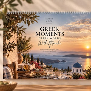 May include: A 2027 wall calendar titled Greek Moments Greek Words With Meraki, featuring a sunset view of a Greek coastal village with a table set for a meal, including olives, bread, and drinks, all under a spiral-bound top edge.