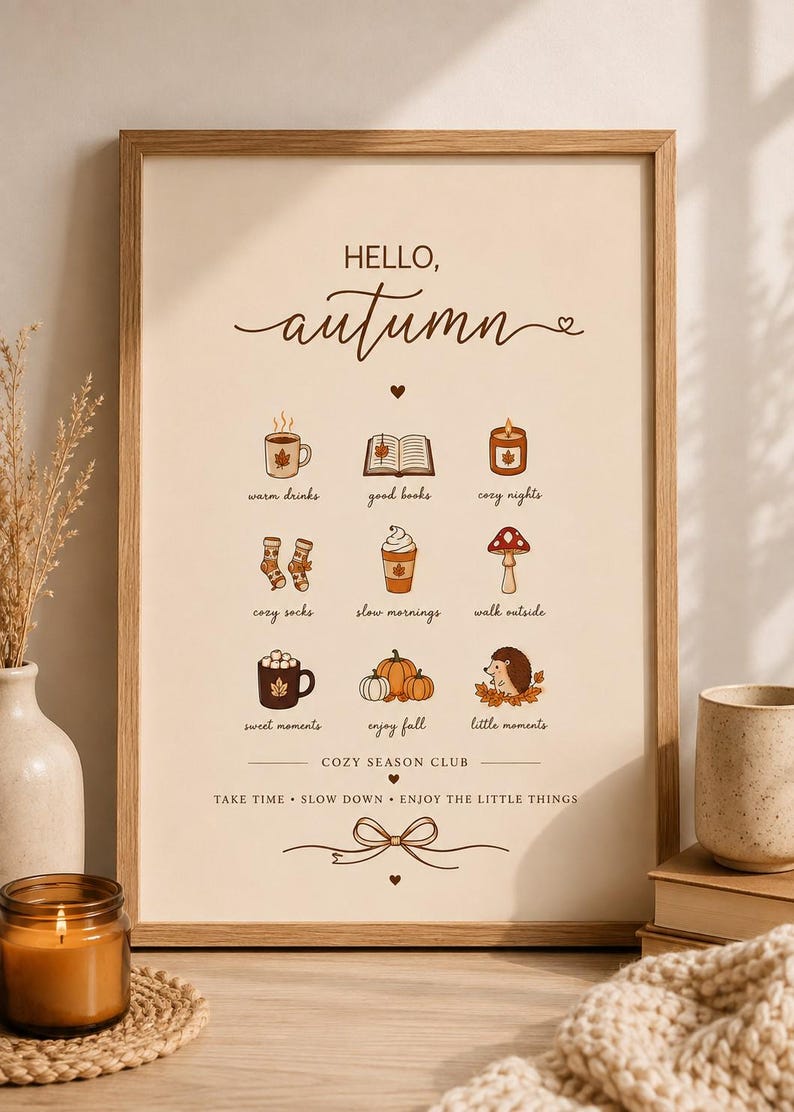 May include: A framed wall art print with a cream background featuring the text HELLO, autumn at the top. Below are nine small illustrations with labels: warm drinks, good books, cozy nights, cozy socks, slow mornings, walk outside, sweet moments, enjoy fall, and little moments. The bottom text reads COZY SEASON CLUB and TAKE TIME, SLOW DOWN, ENJOY THE LITTLE THINGS.