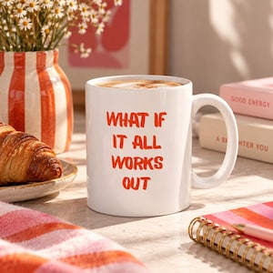 Könnte beinhalten: Ein weißer Keramikbecher mit Henkel, auf dessen Vorderseite der orangefarbene Text "WHAT IF IT ALL WORKS OUT" aufgedruckt ist. Der Becher ist mit einem hellbraunen Getränk gefüllt und steht neben einem Croissant, einer Vase und Büchern.