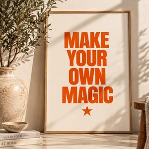 May include: A rectangular framed wall art print featuring the bold orange text MAKE YOUR OWN MAGIC centered on a cream background, with a small orange star icon positioned directly below the final word of the four-line phrase.