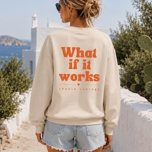 Könnte beinhalten: Ein beigefarbenes Langarm-Sweatshirt mit Rundhalsausschnitt und dem orangefarbenen Schriftzug "What if it works" auf der Rückseite, ergänzt durch ein kleines Herzsymbol über den Worten "choose courage" in einer kleineren, dünneren Schrift.