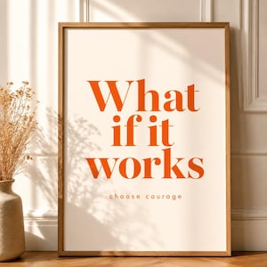 Könnte beinhalten: Ein gerahmter Kunstdruck mit dem orangefarbenen Text What if it works und dem kleineren Text choose courage auf cremefarbenem Hintergrund. Der Druck befindet sich in einem hellbraunen Holzrahmen und zeigt ein klares Typografie-Design.