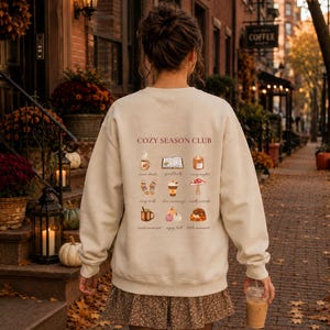 May include: A beige crewneck sweatshirt featuring the text COZY SEASON CLUB and a grid of nine small illustrations including a mug, an open book, a candle, socks, a coffee cup, a mushroom, a copper pot, pumpkins, and a cat, all in warm autumn tones.
