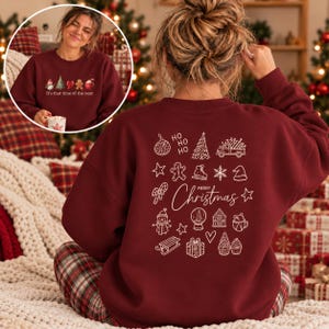 May include: A maroon long-sleeve sweatshirt featuring a white line-art graphic on the back with various holiday icons including a truck, snowman, and gifts, with the text Merry Christmas. The front shows a small colorful holiday graphic.
