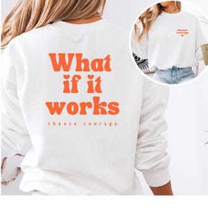 Könnte beinhalten: Ein weißes Langarm-Sweatshirt mit orangefarbenem Text auf der Rückseite, der "What if it works" lautet, darunter "choose courage". Auf der Brust befindet sich der kleinere orangefarbene Text "choose courage" mit einem kleinen Herzsymbol.