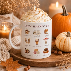 May include: A white ceramic mug filled with whipped cream and topped with cinnamon, featuring the text COZY SEASON CLUB and nine illustrations including a coffee cup, book, candle, socks, drink, mushroom, mug, pumpkins, and a hedgehog.