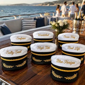 May include: A collection of seven white and black nautical caps featuring gold braided trim and embroidered text including Seafarer, Wanderlust, Ocean King, Salty Soul, High Tide, Reel Life, and Blue Horizon, arranged on a wooden table.
