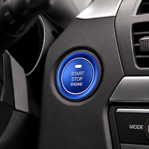 May include: A circular blue engine start stop button installed in a black car dashboard. The button features a metallic blue outer ring and a dark blue center with the white text START STOP ENGINE printed above a small horizontal white indicator light.