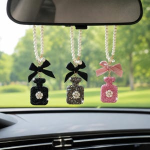 Op de afbeelding: Drie hangende auto-ornamenten in de vorm van parfumflesjes, elk bedekt met sprankelende kristallen. De linker is zwart met een zwarte strik, het midden is grijs met een zwarte strik en de rechter is roze met een roze strik. Elk hangt aan een witte parelketting.