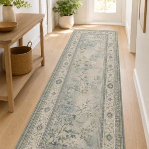 May include: A long rectangular runner rug featuring a faded, distressed pattern in shades of light blue, sage green, and cream. The design includes a central botanical motif with delicate branches and leaves, framed by a decorative floral border.