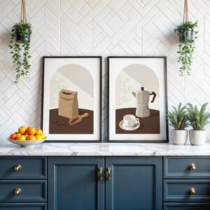 May include: Two framed wall art prints featuring coffee-themed illustrations. The left print shows a brown paper bag of coffee beans and a wooden scoop. The right print displays a silver moka pot and a white coffee cup on a saucer.