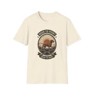 Op de afbeelding: Een crèmekleurig t-shirt met korte mouwen en een ronde afbeelding op de borst. Het ontwerp toont een bruine jachthond in een veld met een zon op de achtergrond, gekruiste geweren en de tekst BORN TO POINT en BRED TO HUNT in het donkergroen.