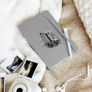 May include: A gray notebook with an elastic closure band and a light blue pen. The cover features a black and white graphic illustration of a city skyline with geometric lines and dots. The notebook is rectangular with rounded corners.