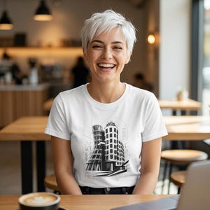 May include: A white short-sleeved t-shirt featuring a black and white architectural line art graphic of a curved building with geometric patterns and structural sketches centered on the chest area, constructed with a crew neckline and standard fit.