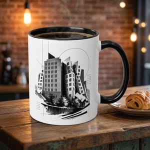 May include: A white ceramic mug with a black handle and rim, featuring a black and white graphic illustration of a cluster of modern buildings with geometric lines, a circular accent, and a dark, textured base design on the front surface.