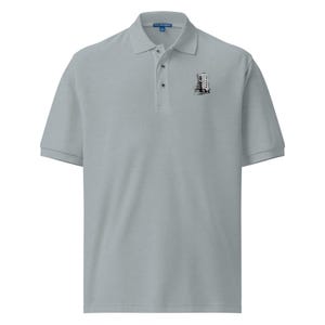 May include: A light gray short-sleeved polo shirt featuring a classic collar, a three-button placket, and a small black and white graphic print on the left chest depicting a stylized architectural building with curved, wavy lines and a dark base.