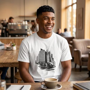 May include: A white short-sleeved t-shirt featuring a black and white graphic print of a cluster of tall buildings with geometric lines and circular accents centered on the chest. The design includes architectural sketches of skyscrapers.