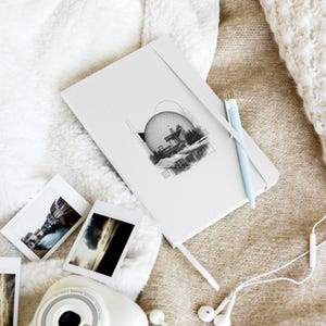 May include: A white hardcover notebook with an elastic closure band features a black and white graphic illustration of a geodesic dome structure. A light blue pen with a gold clip rests on the notebook cover alongside small photo prints.