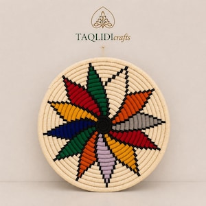 May include: A round, hand-woven wall basket featuring a vibrant, multi-colored starburst pattern in red, green, orange, yellow, blue, grey, and lavender, centered with a black spiral. The text TAQLIDIcrafts appears above the basket.