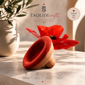 May include: A terracotta-colored ceramic object with a wide, circular, red-painted rim and a tapered base. A vibrant red poppy flower is positioned behind the object. Text on the image includes TAQLIDI crafts, AUTHENTIC HANDMADE MOROCCAN HERITAGE, and COUP DE.