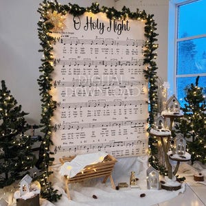 May include: A large vertical sign featuring the O Holy Night musical score and lyrics, framed by a green garland with warm white string lights. A wooden manger with a white cloth sits in front, surrounded by small house ornaments and pine trees.
