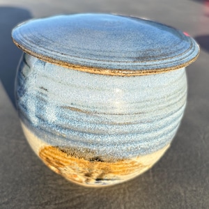 May include: A round ceramic butter keeper with a lid. The upper portion features a textured, mottled blue glaze with horizontal ridges, while the lower base transitions into a smooth, unglazed tan and brown earthy tone with visible speckles.