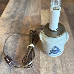 May include: A small electric lamp featuring a speckled off-white ceramic base with two side handles and a blue heart-shaped painted design. The lamp has a brass-colored metal top, a white candle-style socket, a clear light bulb, and a brown cord.