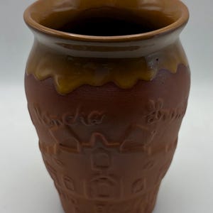 May include: A terracotta-colored ceramic mug with a smooth, glossy brown glaze dripping from the rim. The exterior features embossed architectural details of buildings and houses, along with faint, raised script text integrated into the design.