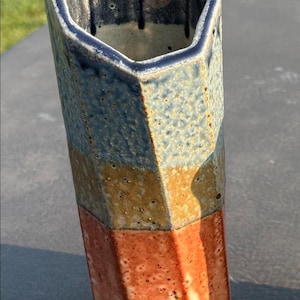 May include: A tall, hexagonal ceramic vase featuring a textured surface with horizontal bands of color. The top section is blue, the middle is a muted mustard yellow, and the bottom is a reddish-orange, all with a speckled, matte finish.