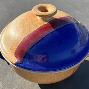 May include: A round ceramic casserole dish with a lid, featuring a speckled tan base and a lid with a glossy deep blue glaze, accented by a diagonal band of reddish-purple. The lid has a central knob handle and the base has side handles.