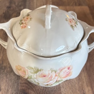 May include: A cream-colored ceramic sugar bowl with a matching lid, featuring a scalloped rim and two side handles. The exterior is decorated with delicate, soft pink rose floral patterns on both the body and the lid, finished with a glossy glaze.