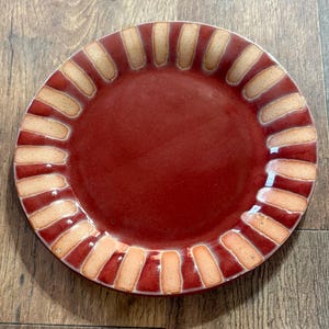 May include: A round ceramic plate with a deep red, glossy center and a wide rim featuring a repeating pattern of alternating dark red and light beige rectangular segments arranged in a radial design around the edge of the plate.