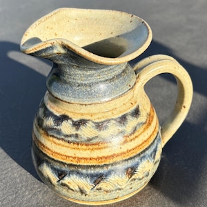 May include: A ceramic pitcher with a wide, flared spout and a curved handle. The body features a speckled beige base with horizontal bands of dark blue and brown, accented by textured, wave-like patterns around the center and base.